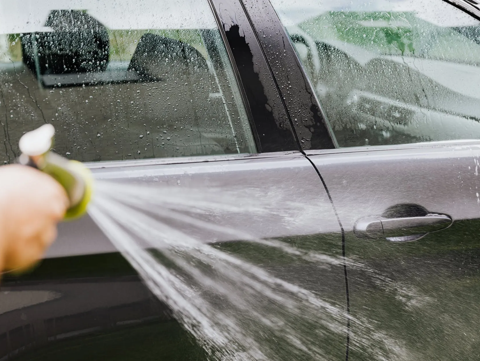 Person spraying a grey car with water, washing it outdoors.