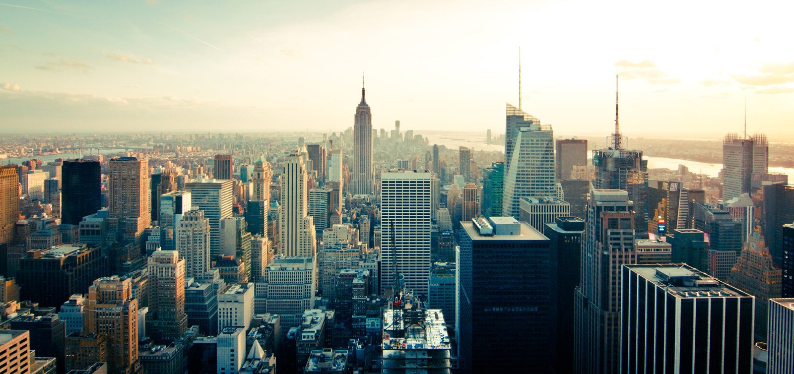Panoramic view of New York City skyline at sunrise, with numerous skyscrapers and a river in the distance.