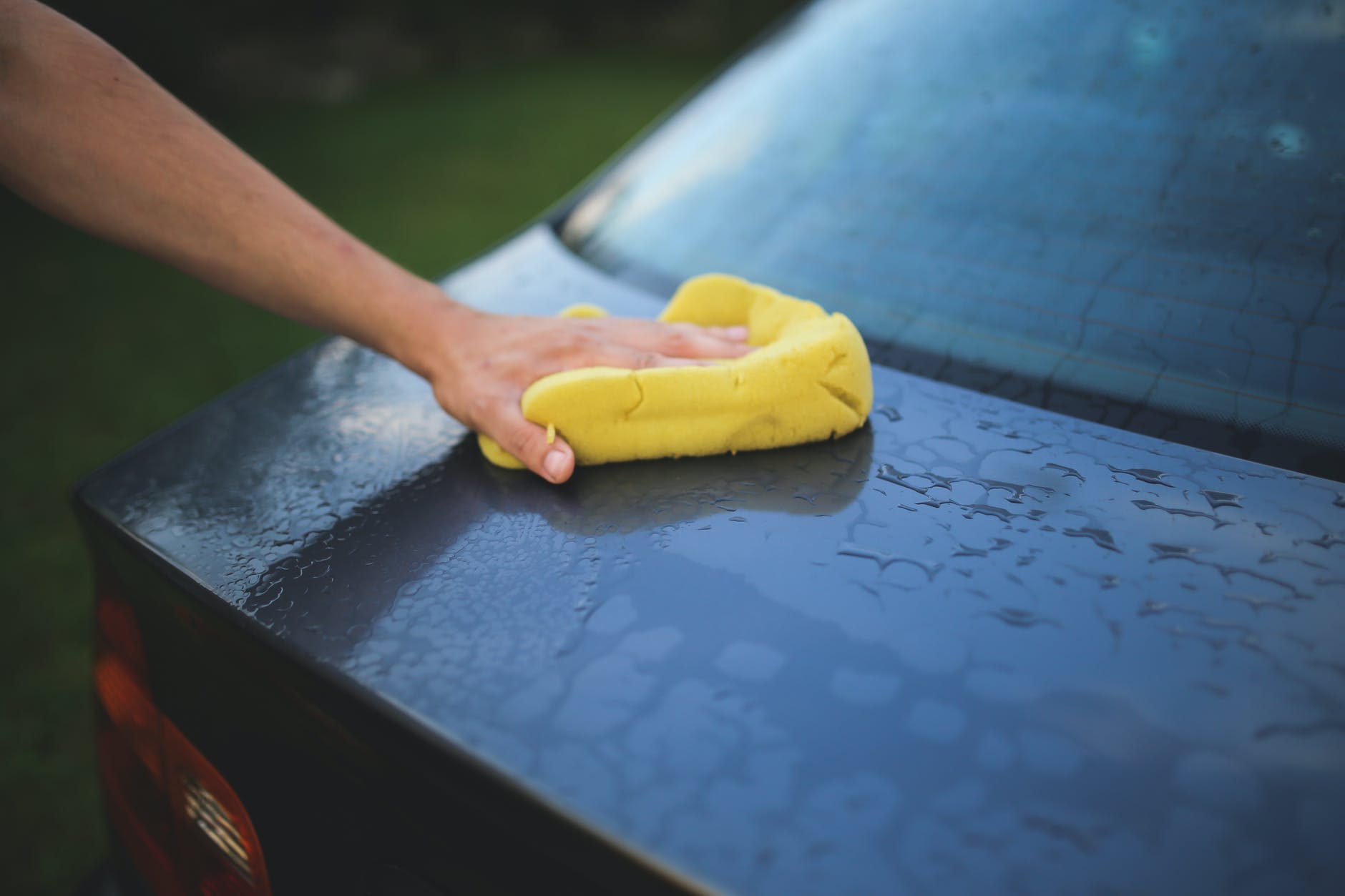 Hand washing a dark blue car with a yellow sponge.